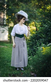 Full-lenght Portrait Of A Young Slender Woman In A 1910s Costume. A Lady In A Hat In The Fashion Of The Early 20th Century Walks In A Garden