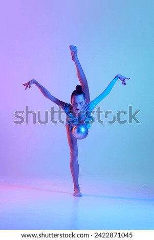 Full-length dynamic image of young talented girl in sparkling costume performing with ball against gradient studio background in neon light. Concept of sport, beauty, competition, art, youth, hobby