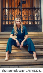 A Full-length Girl Sits On The Stairs In A Long Blue Overalls. The Girl Looks Into The Camera With A Daring Look. Blonde With Long Legs Poses.