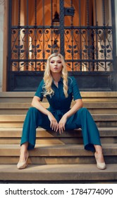 A Full-length Girl Sits On The Stairs In A Long Blue Overalls. The Girl Looks Into The Camera With A Daring Look. Blonde With Long Legs Poses.