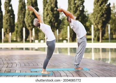 Full-length Of Healthy Elderly Couple Of Husband And Wife In Sportswear Practicing Partner Yoga Outside Against City Park Background, Standing Barefoot On Mats In Mountain Pose With Hands Raised Up