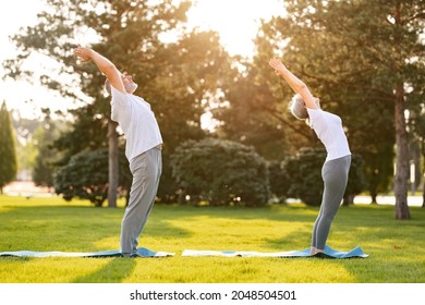 Full-length Of Healthy Elderly Couple Of Husband And Wife In Sportswear Practicing Partner Yoga Outside Against City Park Background, Standing Barefoot On Mats In Mountain Pose With Hands Raised Up