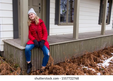 Full-length Horizontal Photo Of Pretty Smiling Young Woman With Long Blond Hair Wearing Red Puffy Winter Coat And Jeans Sitting On Wooden Porch Leaning On Column In Late Fall