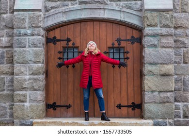 Full-length Horizontal Photo Of Pretty Smiling Young Woman With Long Blond Hair Wearing Red Puffy Winter Coat And Jeans Standing With Arms Wide Open And Head Up In Front Of Old Wooden Door