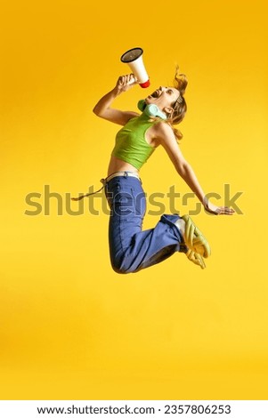 Full-length image of active, positive, young women in stylish clothes jumping with megaphone against yellow studio background. News. Concept of human emotions, youth, fashion, lifestyle, ad