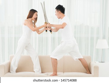 Full-length image of a young couple fighting with swords on the sofa at home