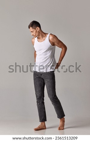 Full-length image of young guy, handsome man posing in singlet and jeans against grey studio background. Concept of men's health and beauty, body care, fitness, wellness, ad