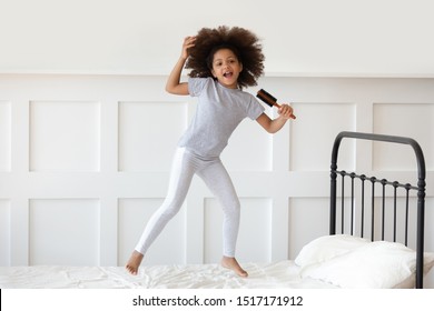 Full-length of little african girl in night-wear awoken jumping on bed holding hair brush like microphone open mouth singing imitates imagines herself a real singer, hobby mood pastime at home concept