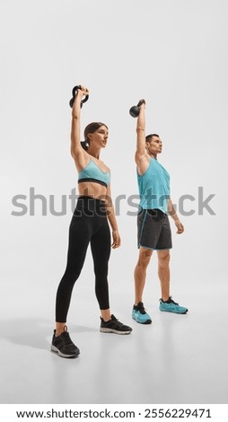 Full-length low angle side view image of young man and woman in sportwear training, doing kettlebell overhead carry exercise against white background. Concept of sport, active and healthy lifestyle