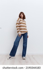 a full-length photo of a beautiful, stylish woman standing on a light background in a striped sweater and blue jeans posing in a relaxed manner