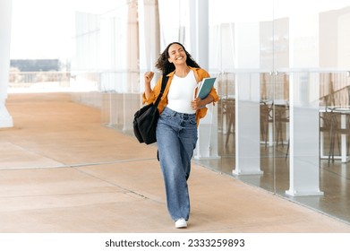 Full-length photo of cheerful amazing curly haired mixed race female student, with a backpack, hold books, notebooks in her hand, walking near the university, looks away and smile, finished school day - Powered by Shutterstock