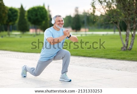 Full-length photo of happy smiling retired sportsman in sportswear and headphones doing front squat on one leg forward with hands folded outside in city park during routine workout in morning
