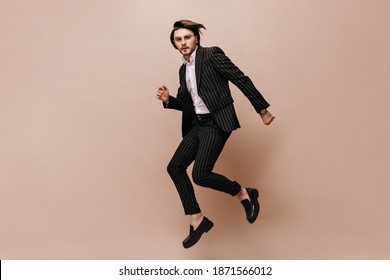 Full-length photo of jumping young man with brunette hair, glasses, white shirt and black suit. Model looking into camera and posing against beige background
