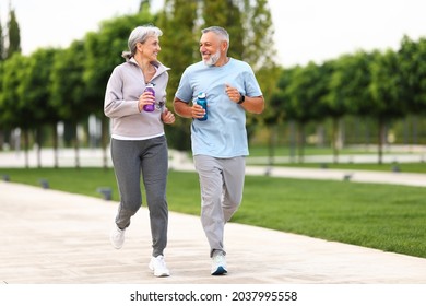 Full-length photo of lovely joyful retirees couple jogging outside in city park along alley with green trees, happy husband and wife looking at each other with smile holding water bottles in hands
