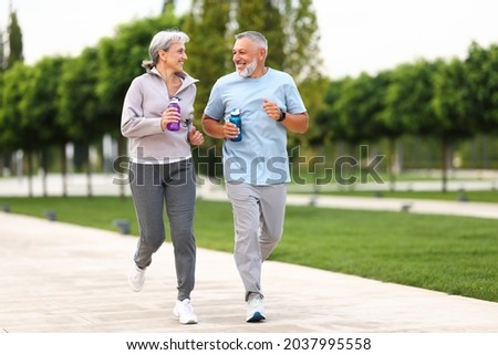 Full-length photo of lovely joyful retirees couple jogging outside in city park along alley with green trees, happy husband and wife looking at each other with smile holding water bottles in hands