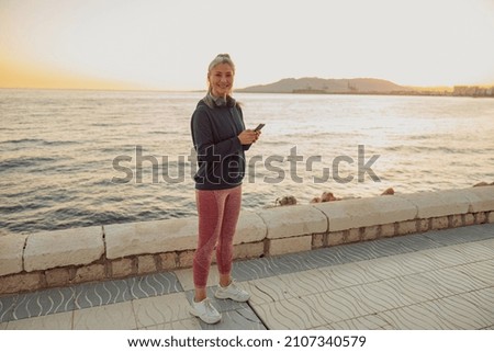 Full-length photo of mature female in sportswear standing on seafront and looking looking at camera on the background of sky at sunset