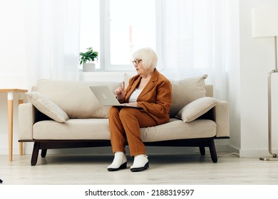 A Full-length Photo Of A Nice Sweet Old Lady Sitting On A Beige Sofa In A Brown Suit Working On A Laptop