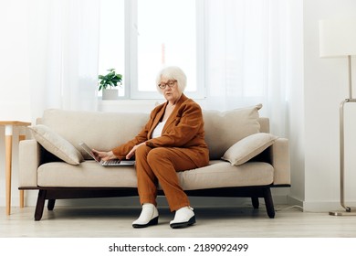 A Full-length Photo Of A Nice Sweet Old Lady Sitting On A Beige Sofa In A Brown Suit Working On A Laptop