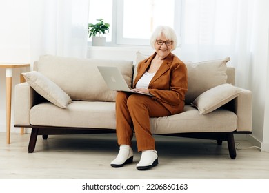 A Full-length Photo Of A Nice Sweet Old Lady Sitting On A Beige Sofa In A Brown Suit Working On A Laptop