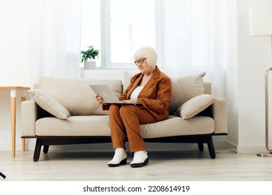 A Full-length Photo Of A Nice Sweet Old Lady Sitting On A Beige Sofa In A Brown Suit Working On A Laptop