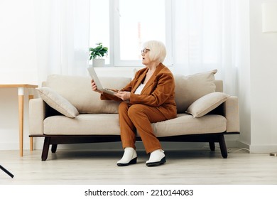 A Full-length Photo Of A Nice Sweet Old Lady Sitting On A Beige Sofa In A Brown Suit Working On A Laptop