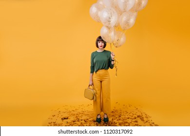 Full-length Photo Of Shy Girl With Short Black Hair Posing In Studio With Party Balloons. Good-looking Caucasian Woman With Elegant Purse Standing On Yellow Background.