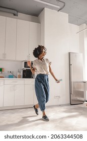 Full-length photo of smiling lady using headphones and dancing on the kitchen while holding smartphone