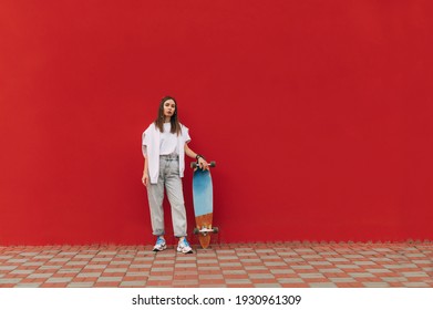 Full-length photo of a stylish skater woman in casual attire standing on a red background, holding her longboard.