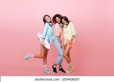 Full-length Photo Of Tanned Sporty Woman With Skateboard Posing In Blue Sneakers Near Friends. Blissful  Lady In Vintage Jeans Relaxing With Girls On Photoshoot In Studio. Place For Text.