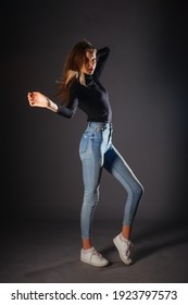 Full-length picture of a beautiful female model posing on dark background in a studio wearing a black blouse, jeans and white sneakers