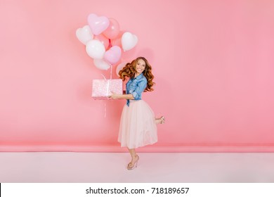 Full-length portrait of adorable laughing birthday girl, jumping with present box and balloons. Gorgeous young woman with curly hair having fun on party holding gift, isolated on pink background.