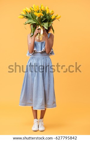 Full-length portrait of african woman in blue dress hiding behind the bouquet of flowers over yellow background