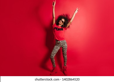 Full-length portrait of amazing black girl posing with hands up. Lovely african lady in sunglasses standing on red background.