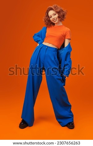 Full-length portrait of attractive joyful fashion model girl with bright red curly hair in blue and orange clothes. Bright colors. Studio orange background. Summer fashion. 