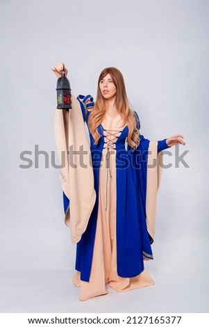 A full-length portrait of an attractive woman with long hair in a medieval, fantasy, blue and beige dress with long, large sleeves, posing isolated on a white background with a lamp in hand.