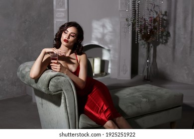 Full-length portrait of a beautiful girl sitting on the couch. Beautiful girl with a glass in her hand. Sexy model in a red dress. Chic makeup. Brunette with beautiful long curls