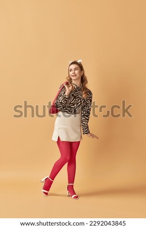 Full-length portrait of beautiful, young, blonde girl in stylish clothes posing against orange studio background. Concept of retro style, fashion, beauty, elegance, 60s, youth. Pin-up style