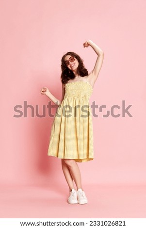 Full-length portrait of beautiful young woman in pretty yellow dress and sunglasses cheerfully dancing on pink studio background. Concept of human emotions, fashion, beauty, lifestyle, summer vibe, ad