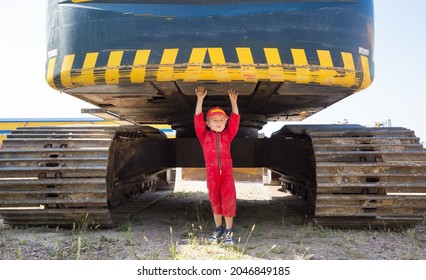 full-length portrait of a boy, a little mechanic, in a red uniform and a cap, standing under a crawler excavator. the boy's hobby for construction equipment, to be like a dad.