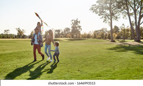 Full-length Portrait Of Cheerful Parents With Two Kids Running With Kite In The Park On A Sunny Day. Family, Kids And Nature Concept. Horizontal Shot.