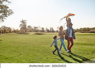 Full-length Portrait Of Cheerful Parents With Two Kids Running With Kite In The Park On A Sunny Day. Family, Kids And Nature Concept. Horizontal Shot.