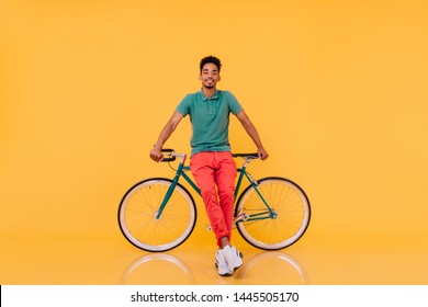 Full-length portrait of confident african man standing in front of his bike. Studio shot of emotional black guy in bright outfit posing with bicycle.