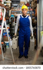 Full-length Portrait Of Confident Hispanic Foreman In Blue Overalls And Hard Hat Carrying Cases With Tools In Building Materials Store