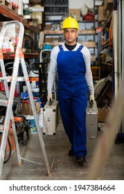 Full-length Portrait Of Confident Hispanic Foreman In Blue Overalls And Hard Hat Carrying Cases With Tools In Building Materials Store