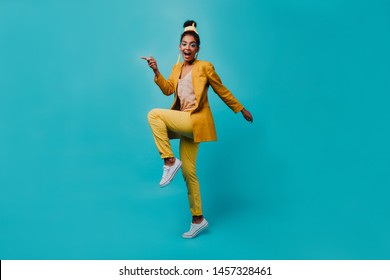 Full-length Portrait Of Fashionable African Woman Jumping On Blue Background. Indoor Photo Of Black Girl Dancing In Yellow Costume.