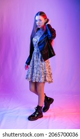 Full-length Portrait Of A Girl. Model In Coarse Boots, Light Chiffon Dress And Leather Jacket. Colored Background. Blue, Red, Purple Light On Background And Person