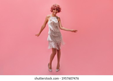 Full-length portrait of good-humored curly pink-haired woman dancing on isolated. Attractive girl in shiny silver dress and high heels smiles on pink background. - Powered by Shutterstock