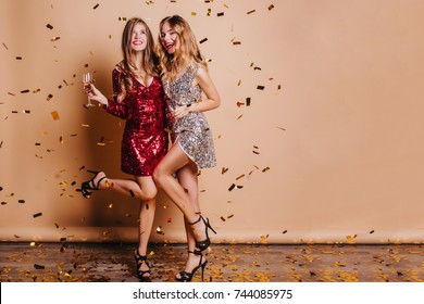 Full-length portrait of happy girls wears high heel shoes dancing at party under confetti. Indoor photo of funny ladies chilling at home during christmas celebration and drinking champagne.