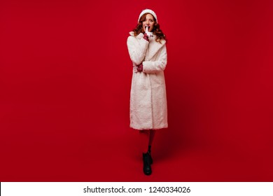 Full-length Portrait Of Magnificent Ginger Woman Standing On Red Background. Refined Female Model In Coat Enjoying Winter Photoshoot.