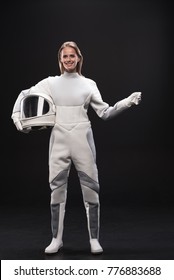 Full-length portrait of optimistic young charming lady cosmonaut is standing in protective suit and holding helmet while gesturing like holding something. She is looking at camera with joy. Isolated